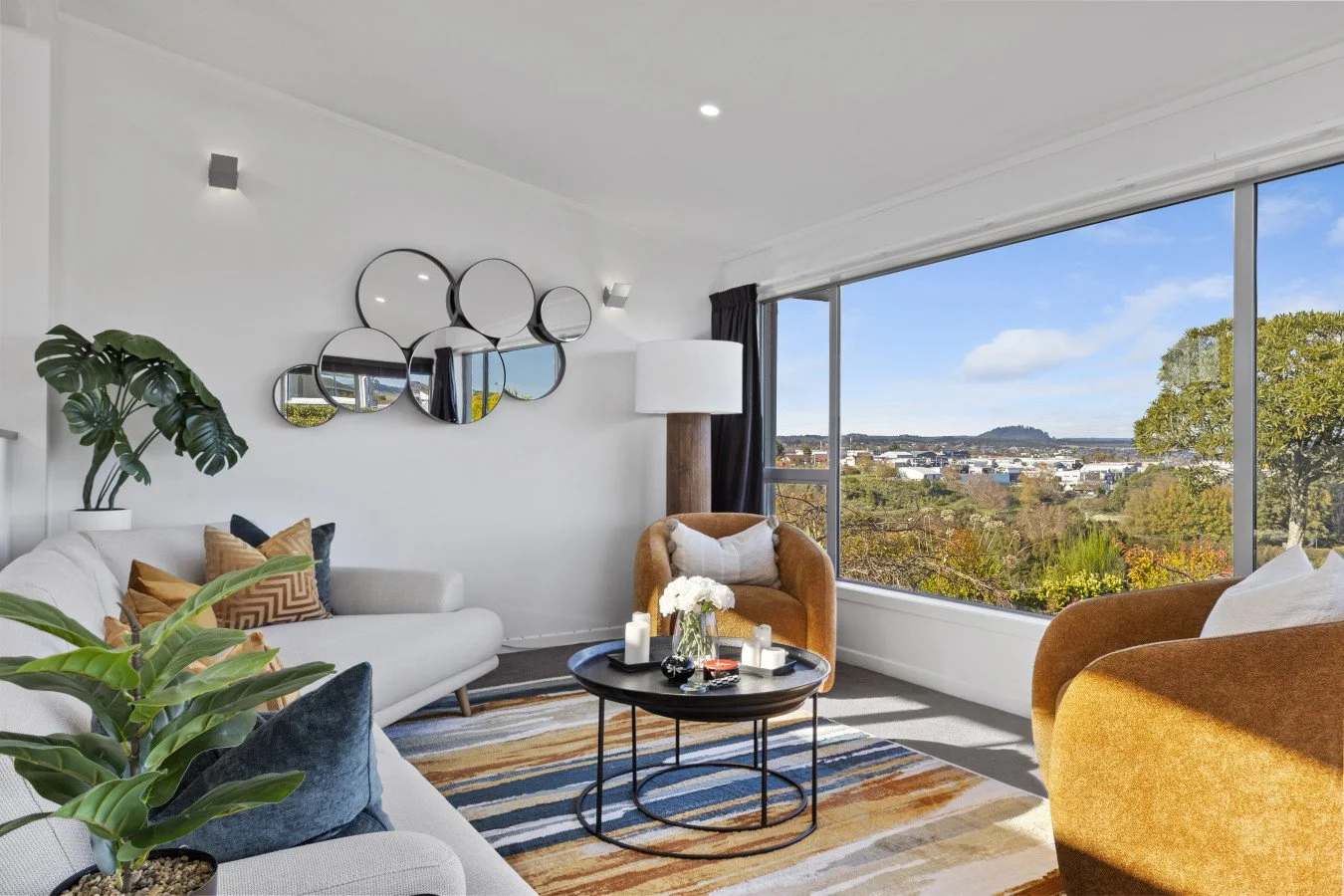Living room with large window showing a scenic view of trees and hills, white sofa with patterned pillows, brown and tan armchairs, a round black coffee table with candles and flowers, a tall white lamp, and modern circular wall mirrors.
