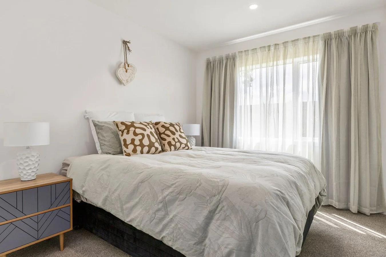 A bedroom with a large bed with white bedding, decorative pillows, a wooden nightstand with a white lamp, and light-colored curtains letting in natural light.