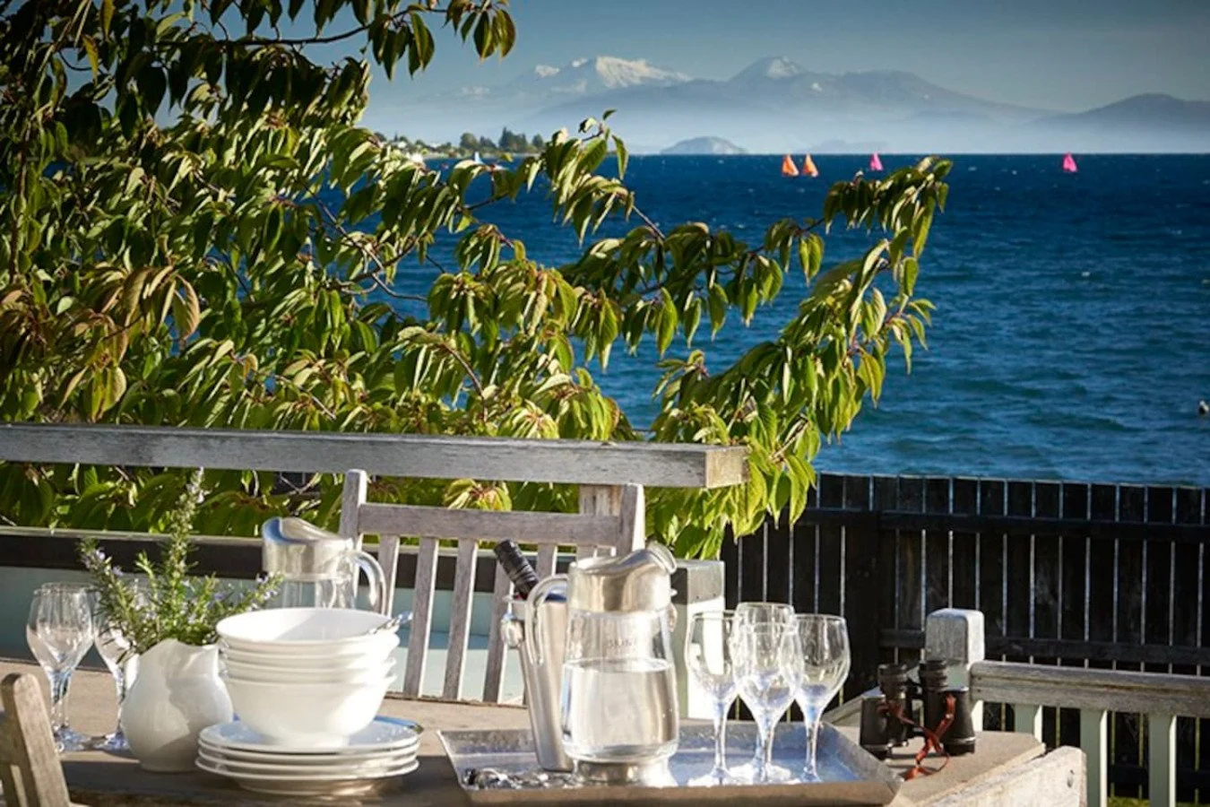 Outdoor dining table set with white plates, glasses, water pitcher, and a small plant, overlooking a lake with sailboats and a mountain range in the background.