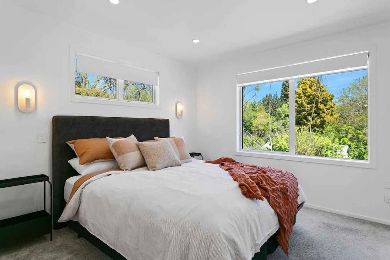 A bedroom with a bed, pillows, and a orange throw blanket, large windows showing green trees outside, and white walls.