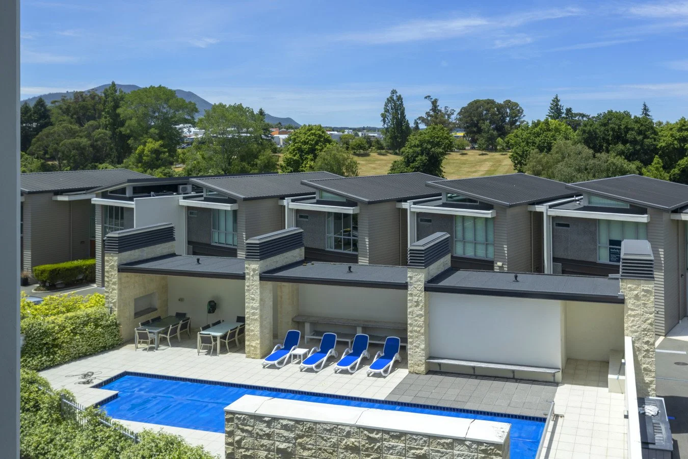 Modern apartment complex with outdoor swimming pool, lounge chairs, and patio tables, surrounded by green trees and hills in the background under a blue sky.