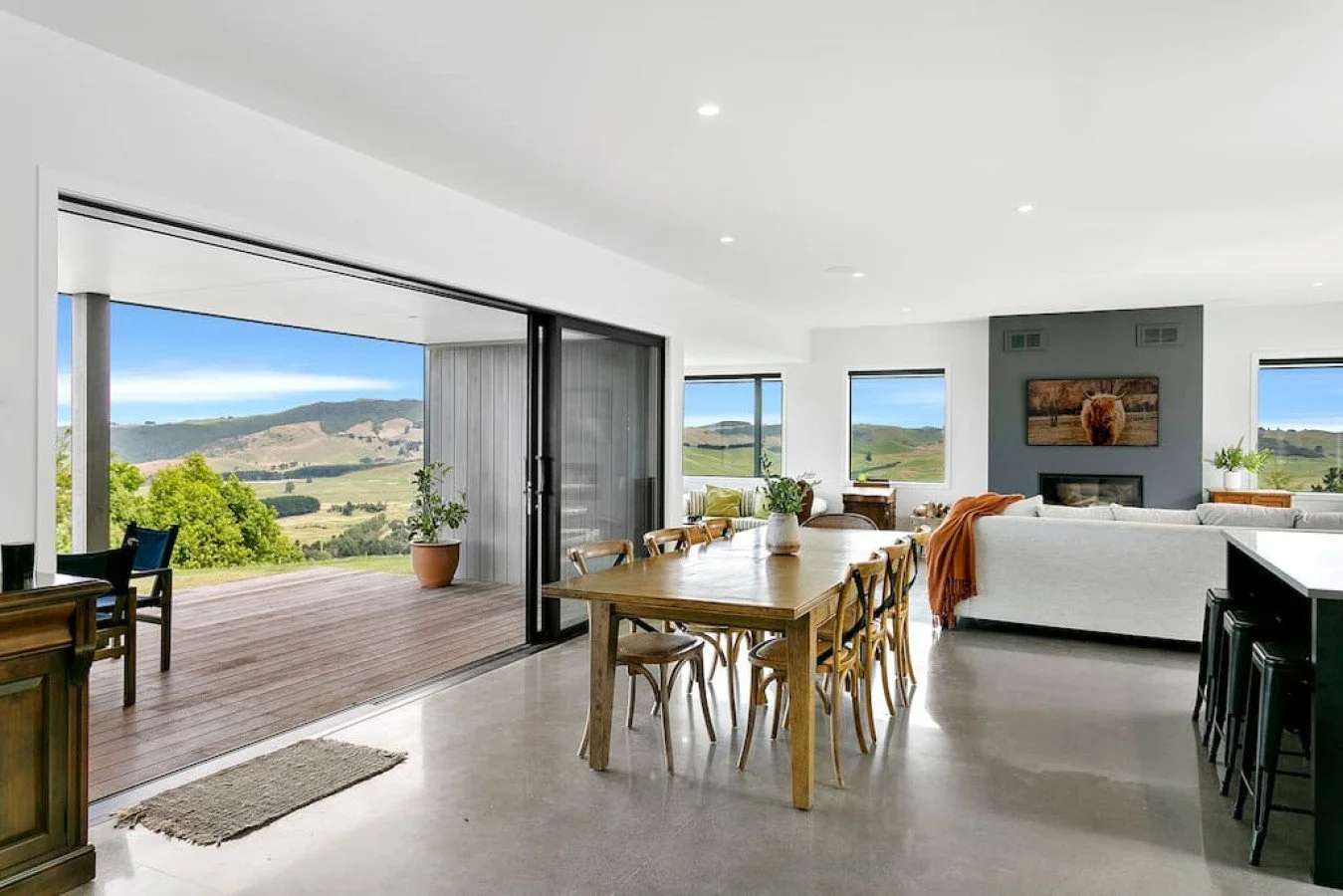 Open-concept living and dining room with large windows showcasing a scenic landscape with hills and blue sky, modern white sofas, a wooden dining table with chairs, and a sliding glass door leading to an outdoor deck with potted plants.