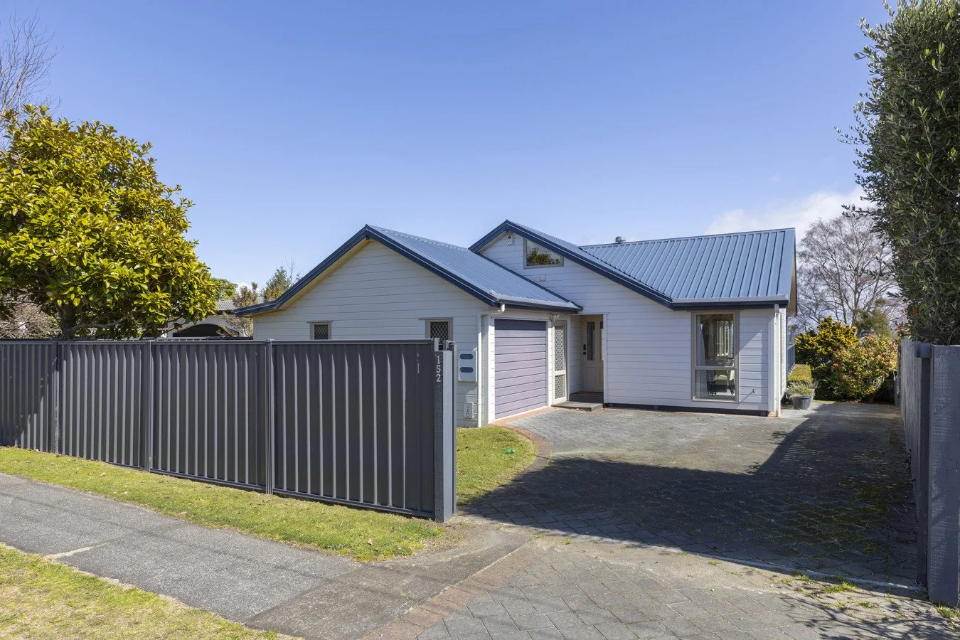 A modern, white house with a blue metal roof surrounded by a gray metal fence, with a driveway and small front yard on a sunny day.