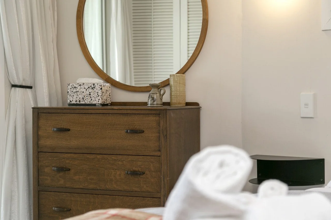 A wooden dresser with a mirror, decorated with a speckled tissue box, a ceramic pitcher, and a woven basket, in a room with white curtains and a white wall.