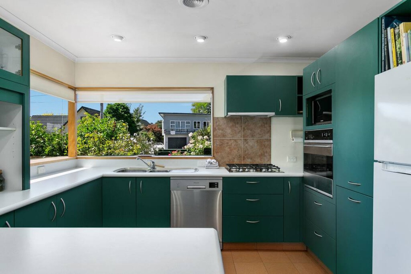 Kitchen with green cabinets, white countertops, a window with a view of houses and trees, and kitchen appliances.