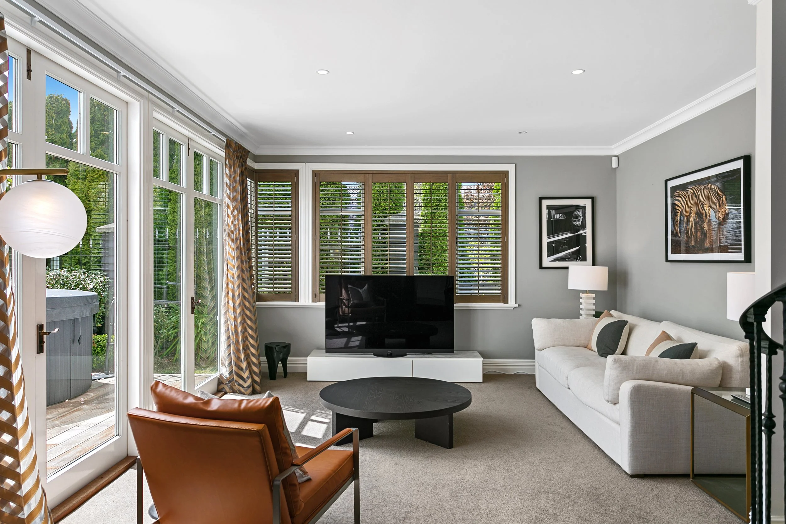 A modern living room with large windows, a white sofa with pillows, a black TV on a white stand, a round black coffee table, a leather armchair, and framed artwork on gray walls.