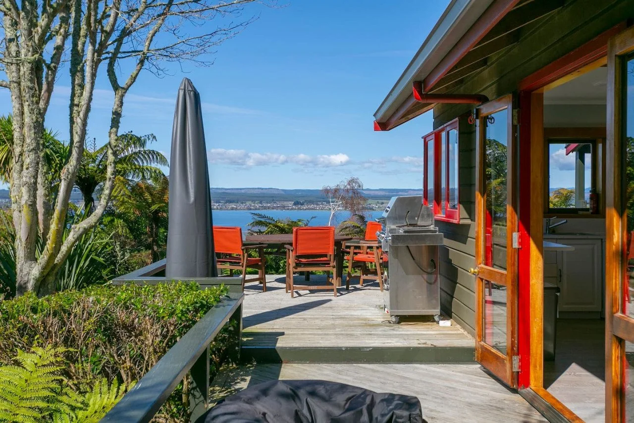 View of a wooden deck outside a house with a barbecue grill, outdoor dining table with chairs, umbrella, surrounded by greenery, overlooking a body of water in the distance.