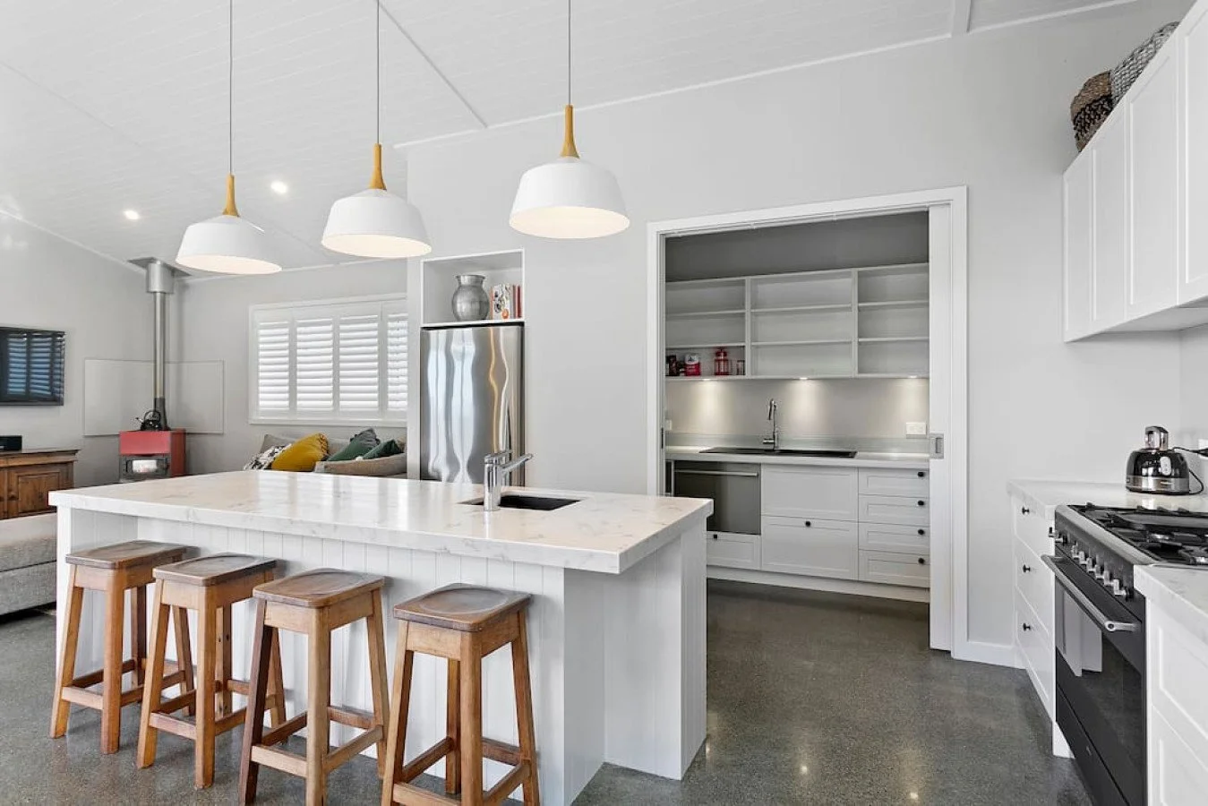 Modern white kitchen with an island counter, wooden bar stools, stainless steel appliances, and pendant lighting.