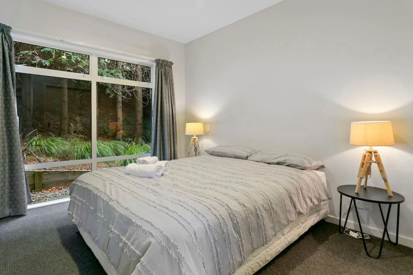 A neatly made bed with white bedding and two pillows in a bedroom with a large window showing trees outside. There are two table lamps on either side of the bed, with one on a black side table. Towels are rolled on the bed.