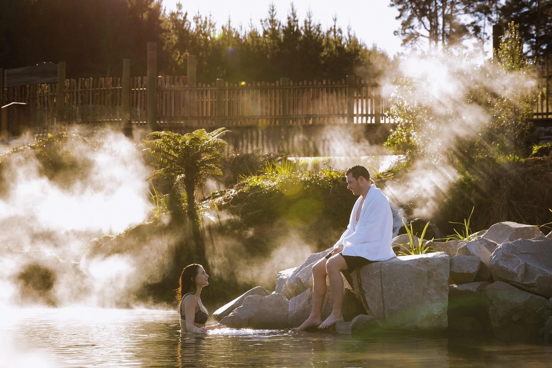 A couple enjoying a steam bath in a hot spring outdoors during sunrise or sunset, with steam rising around them and a scenic background of trees and a wooden fence.