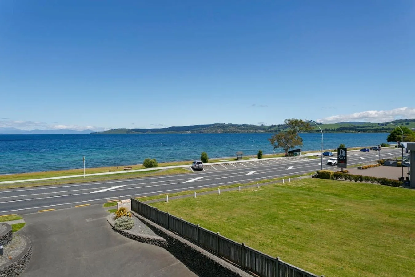 A view of a coastal road next to a body of water with distant hills under a clear blue sky. There's a parking lot with some cars and a grassy area with small plants in the foreground.