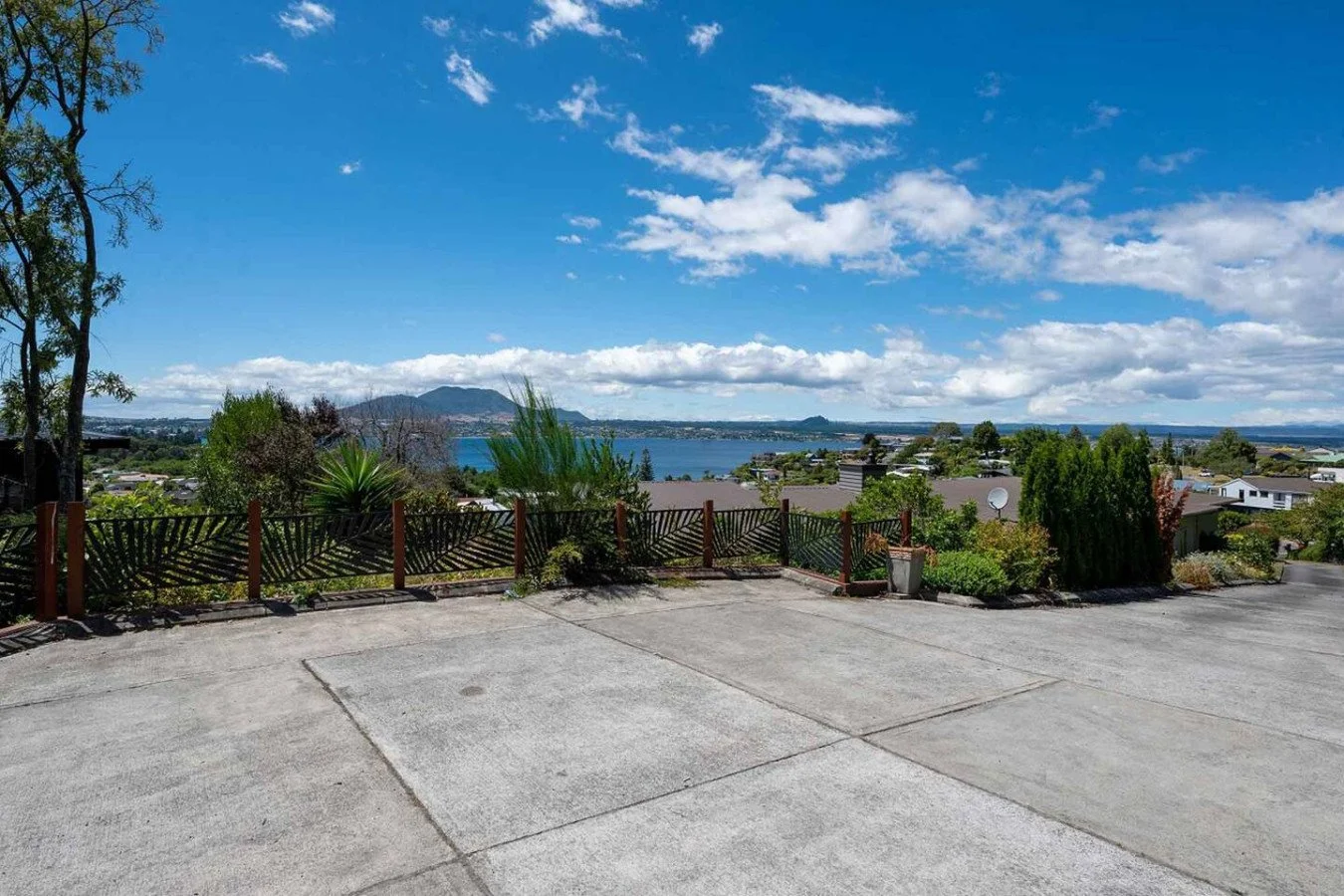 View of a lake with mountains in the background, partly cloudy sky, greenery, and residential houses from a concrete patio with a decorative wooden fence.