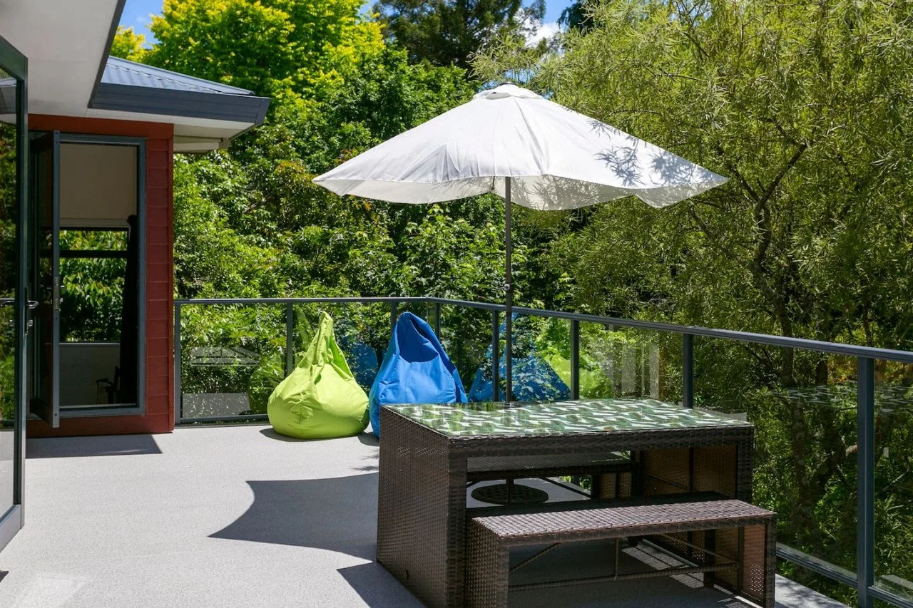 Balcony with white umbrella, two colorful bean bag chairs, and a wicker table with a glass top, overlooking trees.