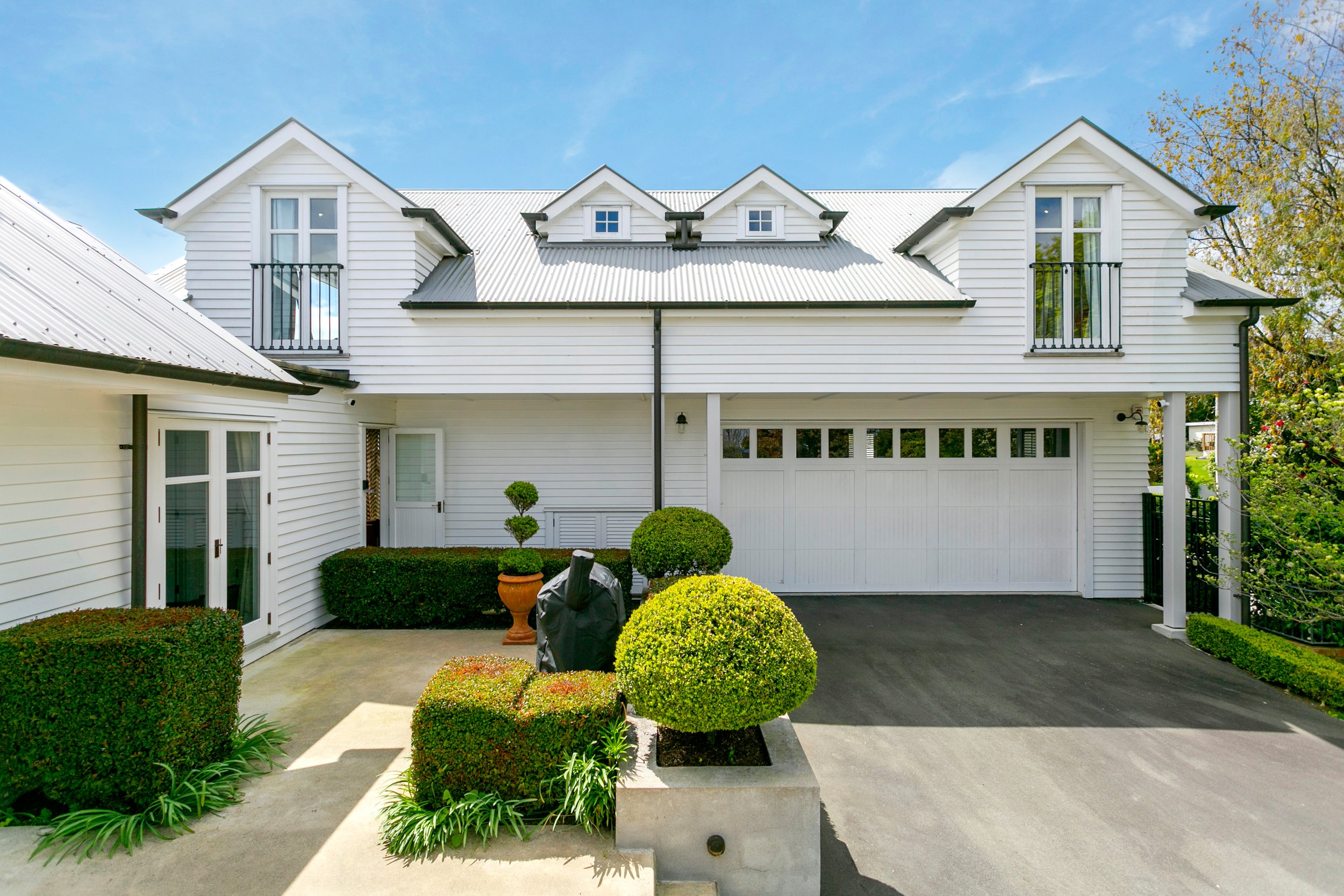 White modern house with gabled roof, black balconies, and a garage, surrounded by neatly trimmed bushes and a concrete driveway.