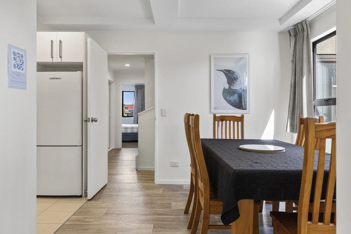 Dining room with wooden table and chairs, black tablecloth, framed bird print on white wall, window with curtains, and open doorway leading to bedroom with window and bed.