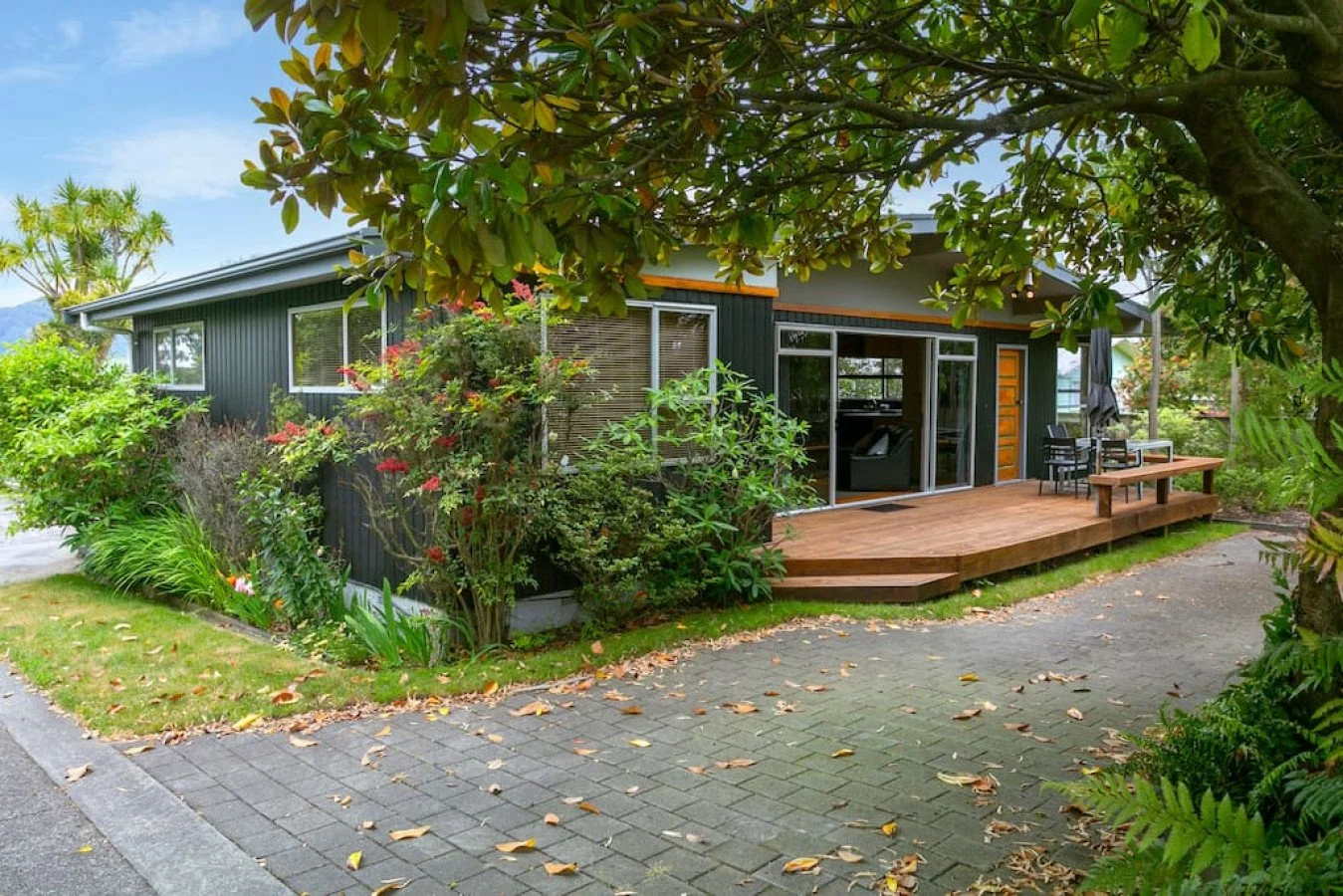Exterior view of a modern house featuring a wooden deck with outdoor furniture, surrounded by lush green plants and trees, with a paved pathway in the foreground.