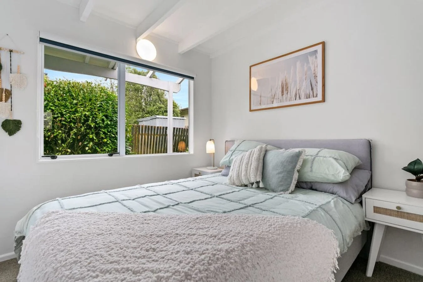 Bright bedroom with a large window overlooking greenery, a neatly made bed with soft pastel bedding, pillows, and a nightstand with a lamp and potted plant.