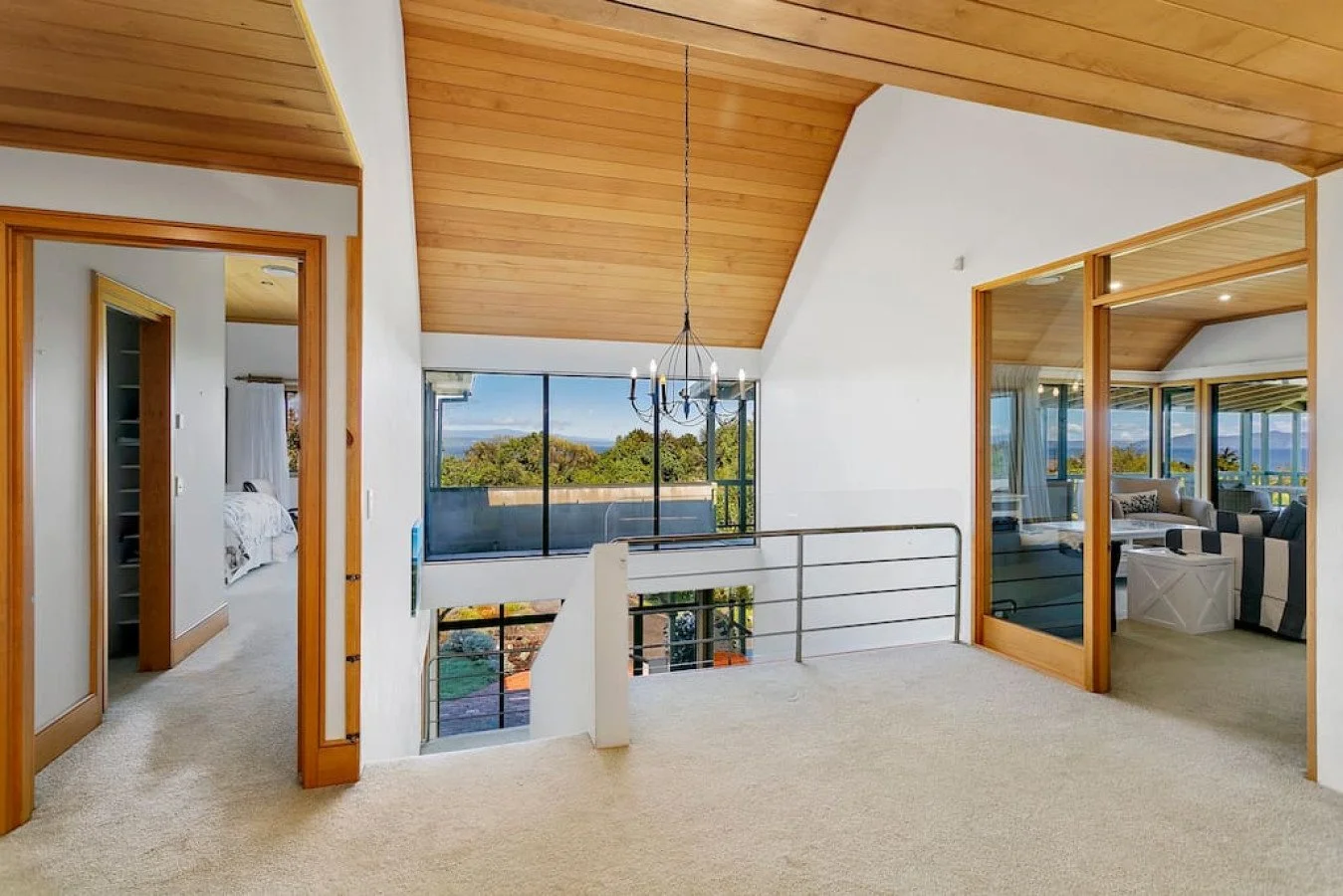 Interior view of a house with a wooden ceiling, large windows, and a chandelier, overlooking a lush green landscape.