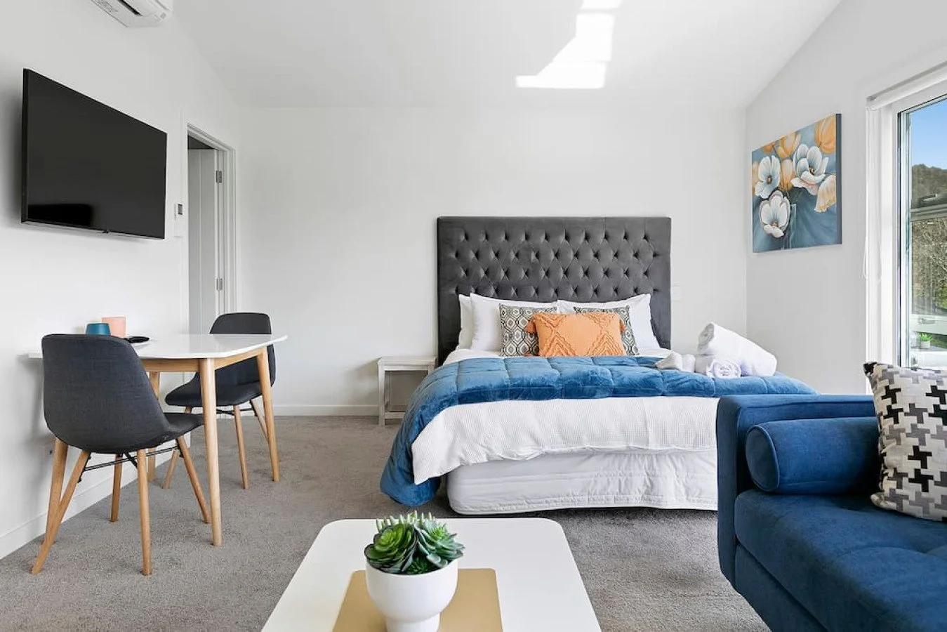 Modern bedroom with a bed, a blue couch, a wall-mounted TV, a small dining table with two chairs, a painting of flowers, and a window letting in natural light.