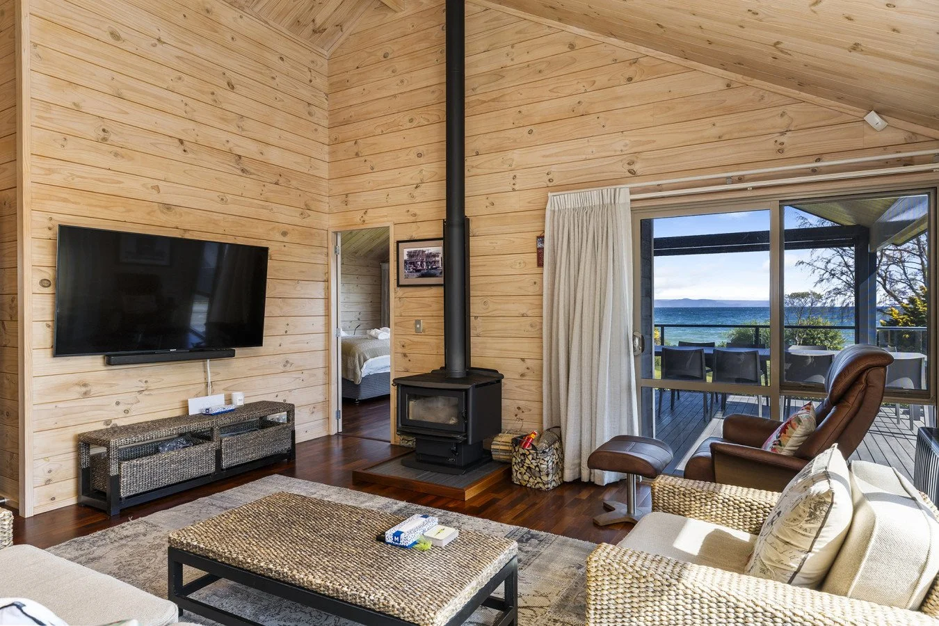 Living room with a wall-mounted TV, a wood stove, a beige sofa, a leather recliner, and large sliding glass doors offering a view of a deck and the ocean beyond.