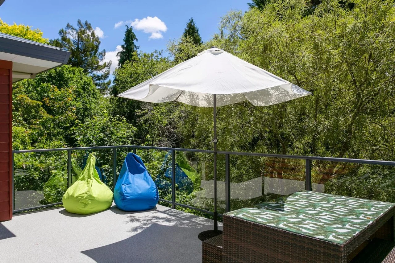 A balcony with a white patio umbrella, two colorful bean bag chairs (lime green and blue), and a wicker lounge chair with a green and white patterned cushion, overlooking lush green trees and a blue sky with scattered clouds.