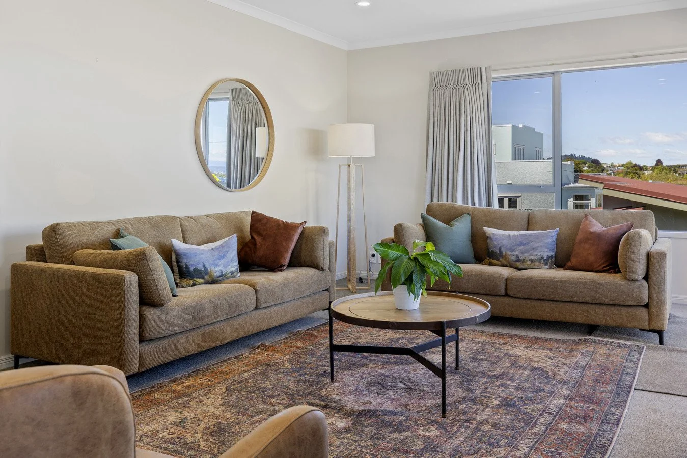 Living room with two beige sofas, throw pillows, a wooden coffee table with a plant, a round mirror on the wall, a tall floor lamp, patterned rug, and a window with curtains showing buildings and a blue sky outside.