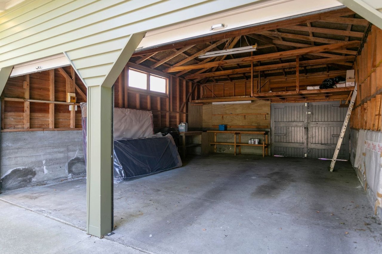 Empty garage with open door, wooden walls, and concrete floor, containing stored items like a ladders, plastic-covered objects, and a workbench.
