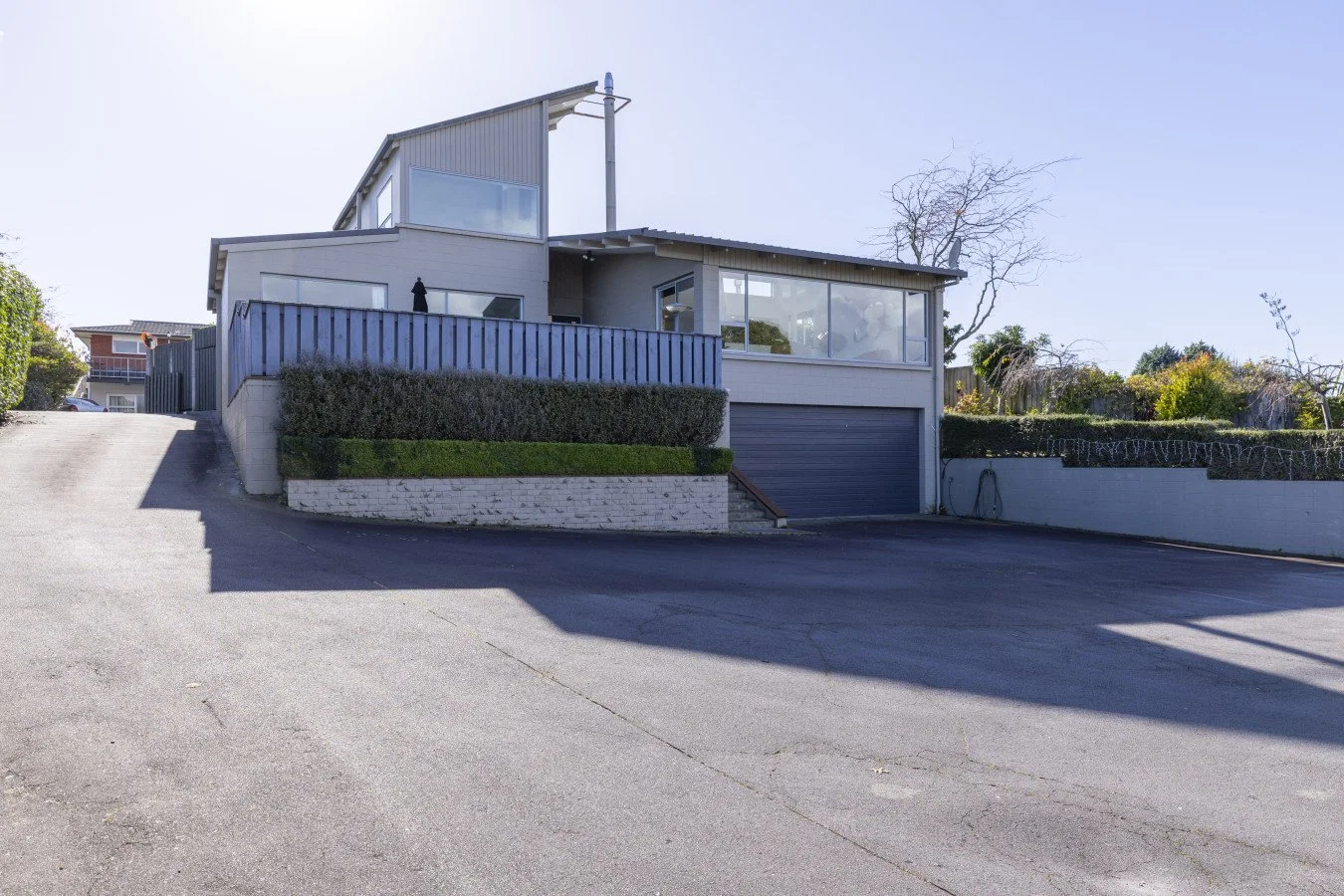 Modern house with multiple levels, large windows, and a garage, situated on a sloped driveway with landscaped bushes and trees.