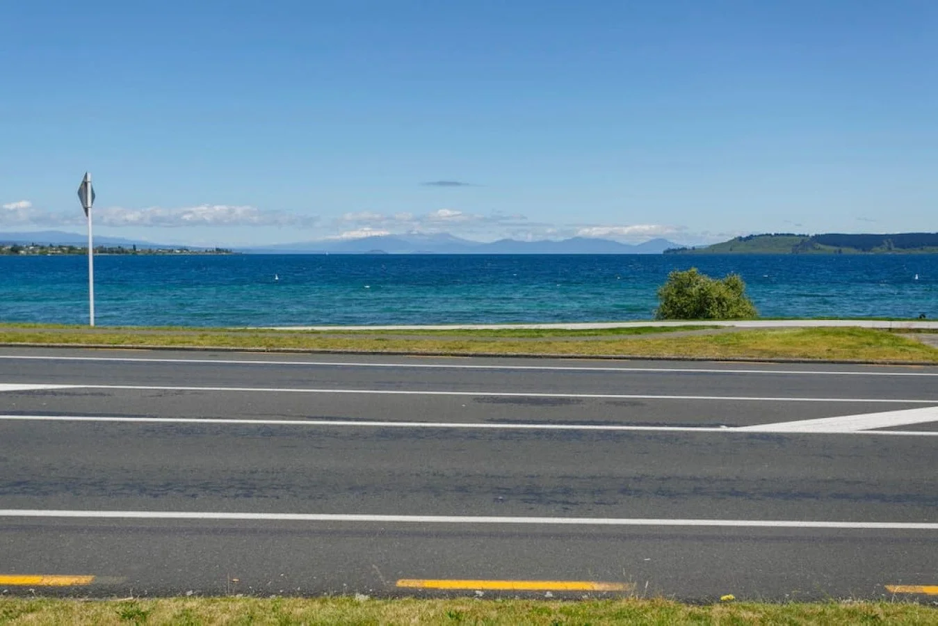 A scenic view of a body of water with a paved road in the foreground, a green bush on the right, and a distant shoreline with hills under a partly cloudy sky.