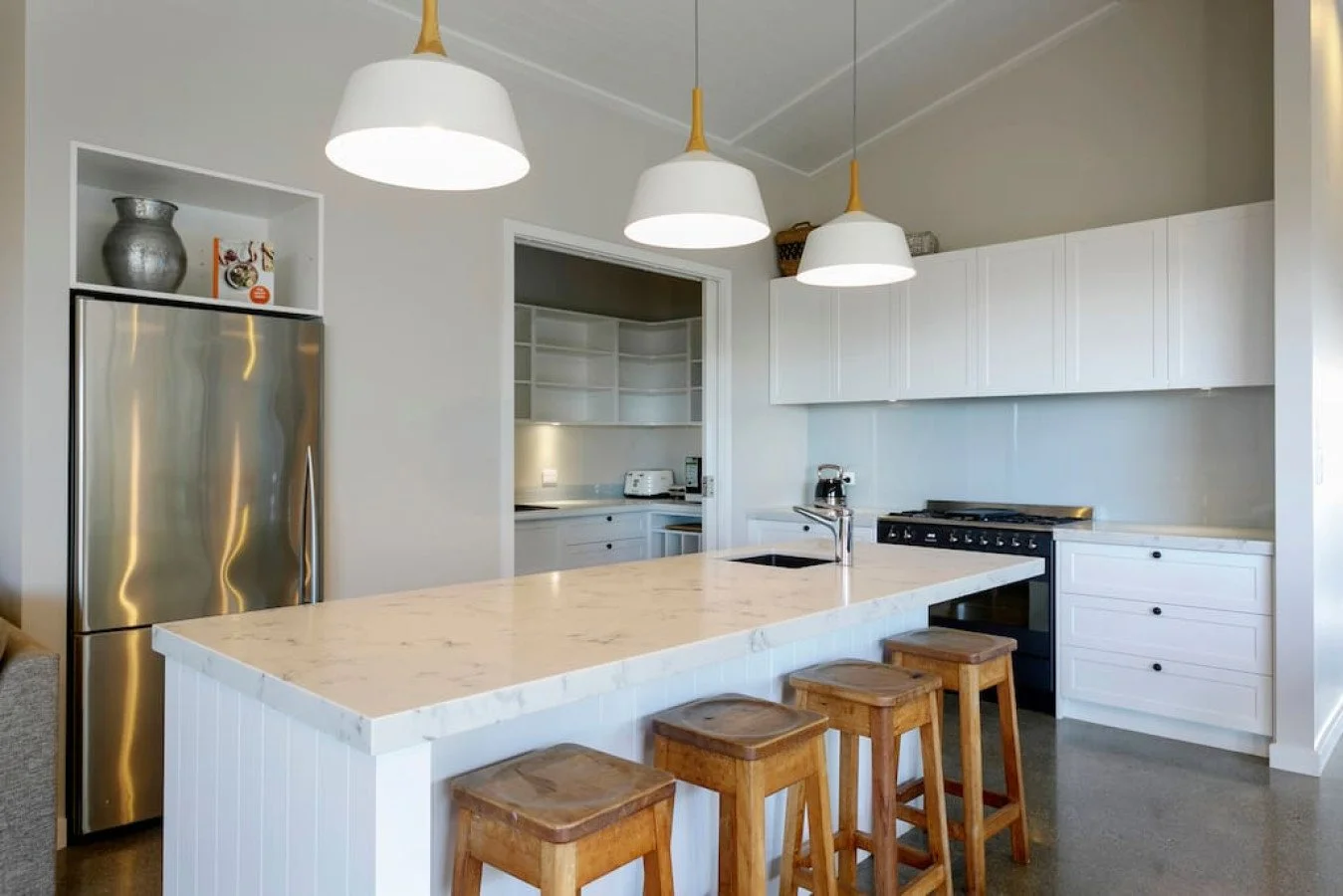 Modern kitchen with white cabinets, stainless steel refrigerator, marble island with four wooden stools, and three white pendant lights hanging from the ceiling.
