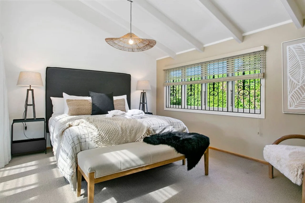 A bedroom with a large bed, black headboard, beige and white bedding, and four pillows. There are two bedside tables with lamps, a window with blinds and iron security bars, a bench at the foot of the bed with a black fur throw, and a framed wall art