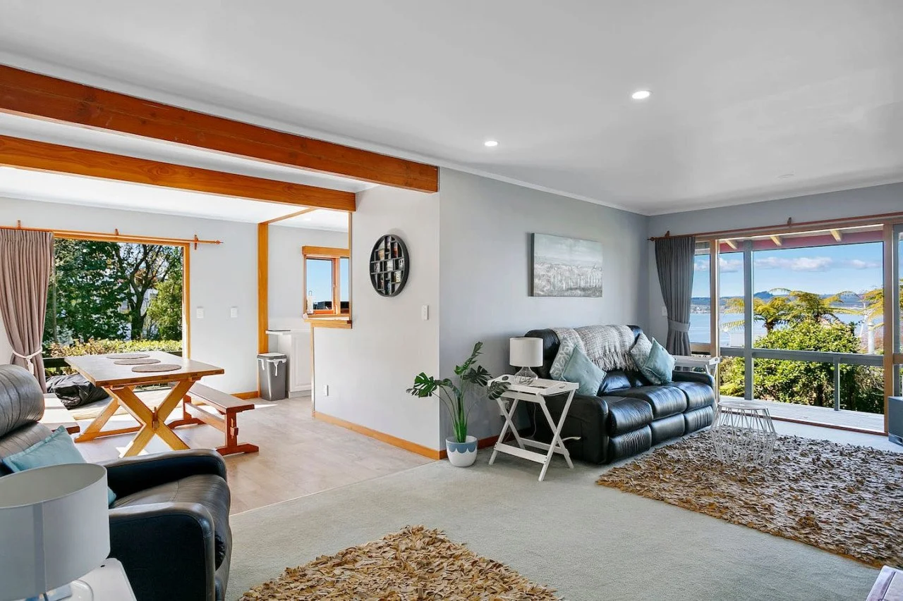 Bright living room with sliding glass doors overlooking water and trees, black leather sofa with light blue pillows, white side table with lamp, houseplant, and a dining area with wooden table and benches.
