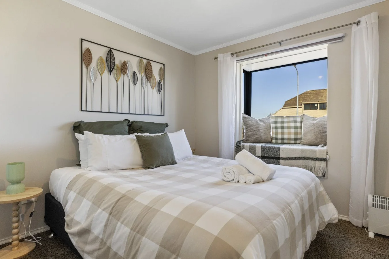 A cozy bedroom with a neatly made bed featuring white and gray checkered bedding, green and gray pillows, and a rolled towel on top. A small wooden nightstand with a green lamp is beside the bed. There is a window with white curtains and a cozy windo