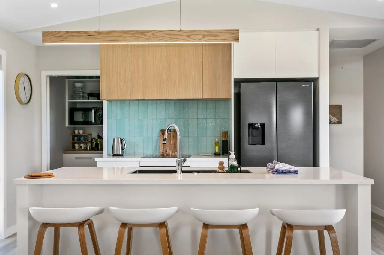 Modern kitchen with a white island counter, four white and wooden barstools, a stainless steel sink, a wooden cutting board, a teal tile backsplash, and a black Samsung refrigerator with a water and ice dispenser.