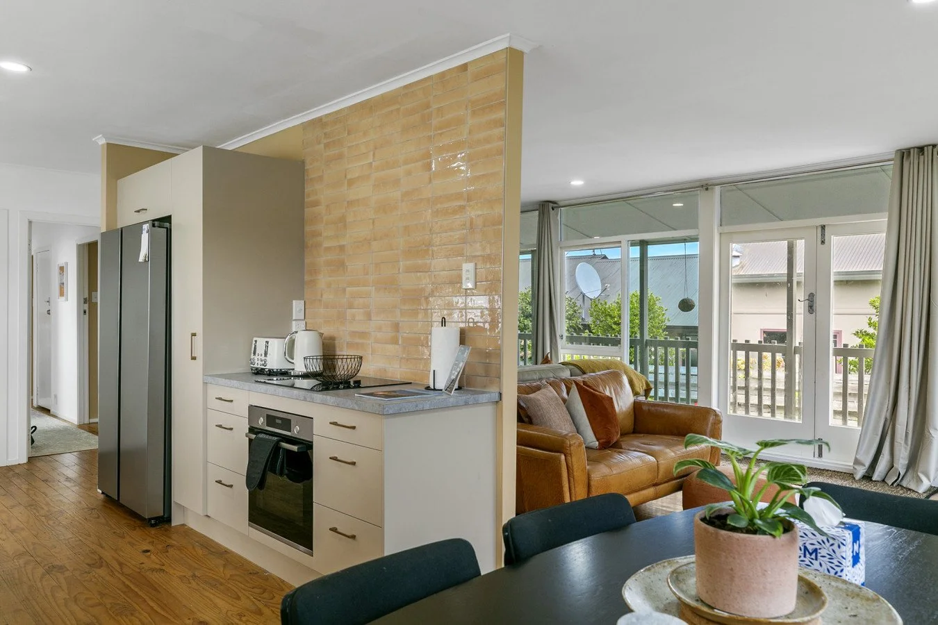 Open-plan living room and kitchen with large windows, beige brick wall, leather sofa, dining table with plant, and kitchen appliances.