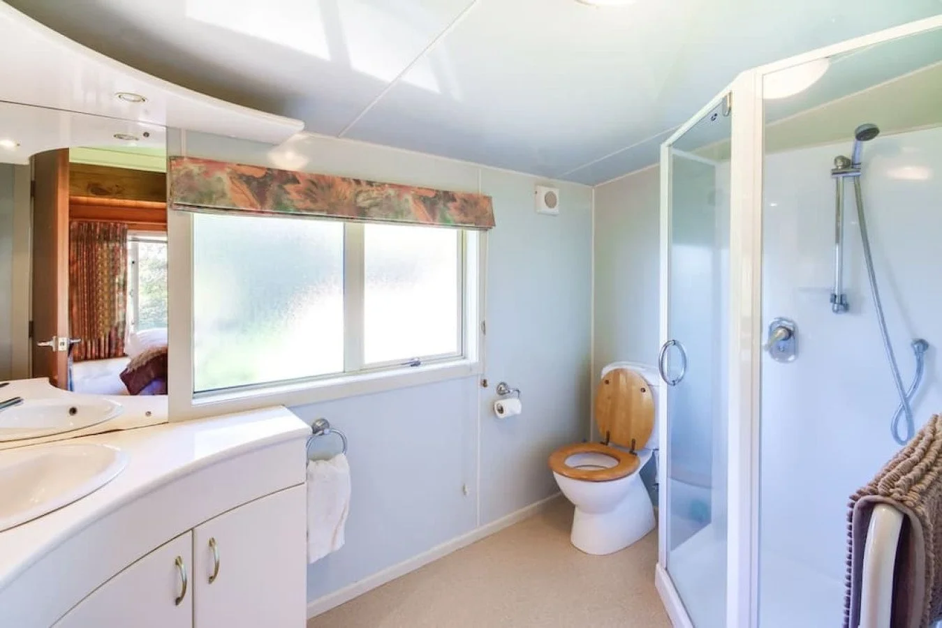 A small bathroom with a window above a white sink and cabinet, a toilet with a wooden seat cover, and a glass shower enclosure with a handheld showerhead.