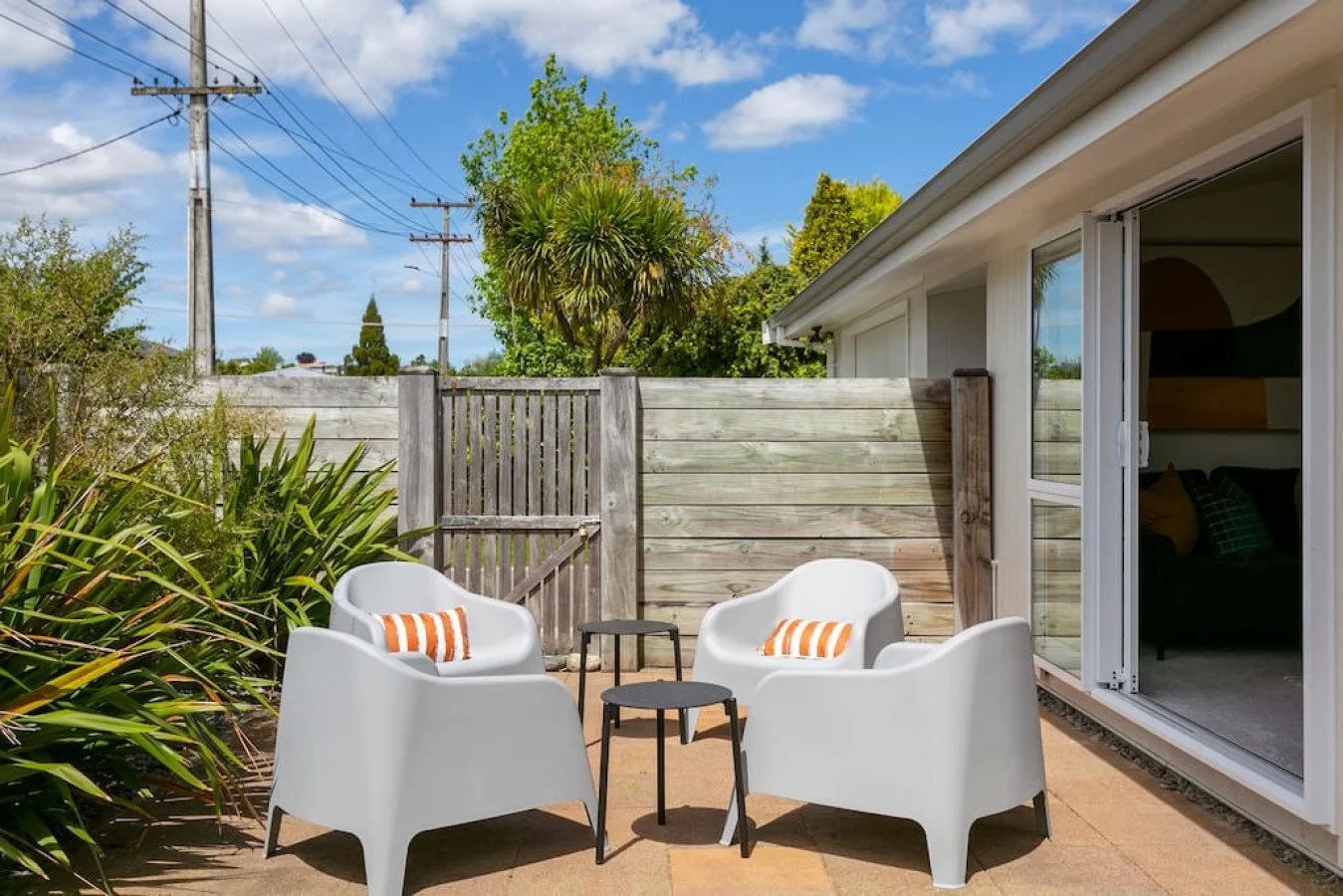 Outdoor seating area with four white chairs, small black tables, orange and white striped pillows, wooden fence, and green plants, adjacent to a house with sliding glass door, under a blue sky with some clouds.