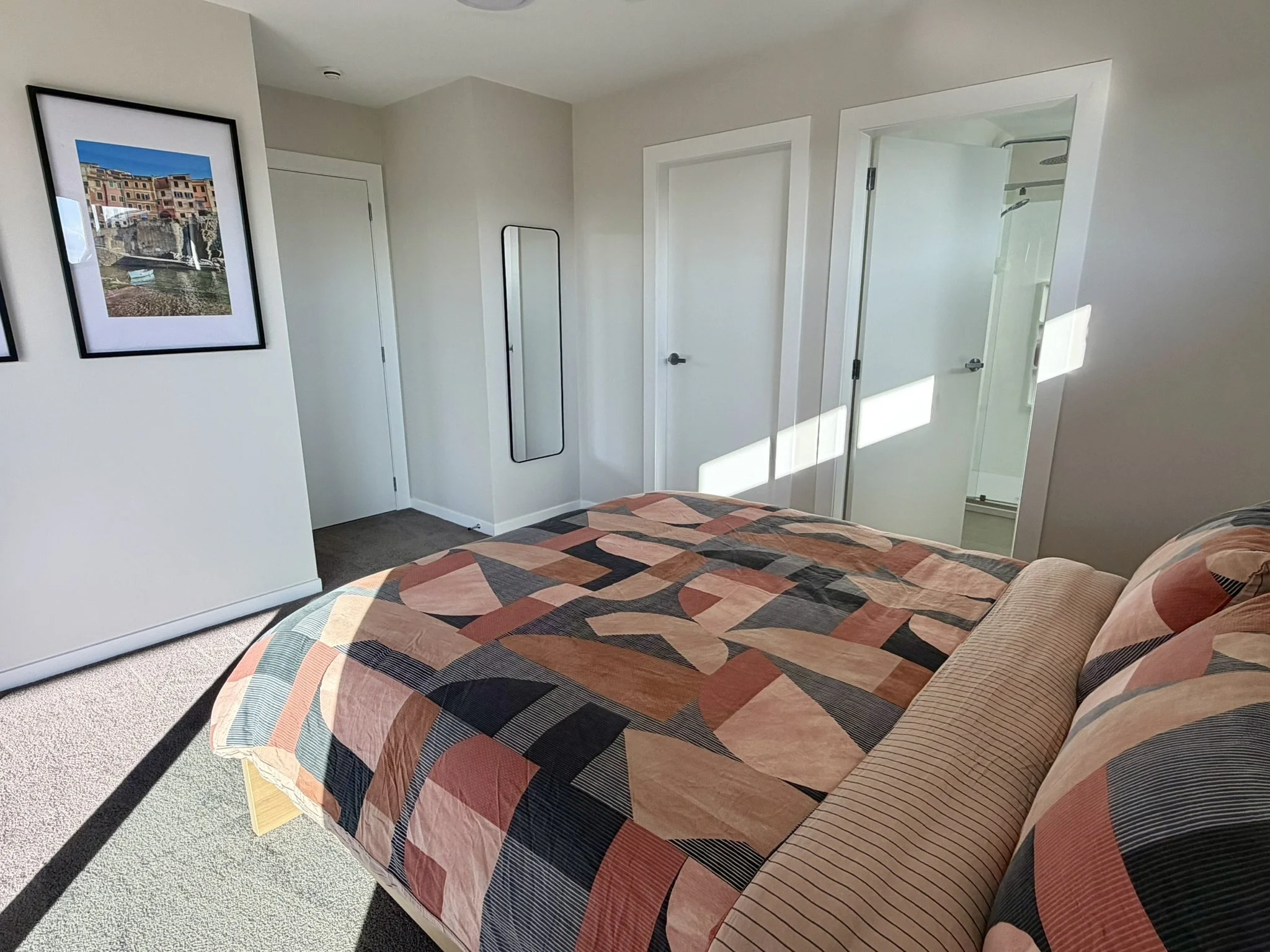 Bedroom with a bed covered in a geometric patterned quilt, a mirror on the wall, three white doors, framed artwork on the wall, and sunlight streaming in through a window.