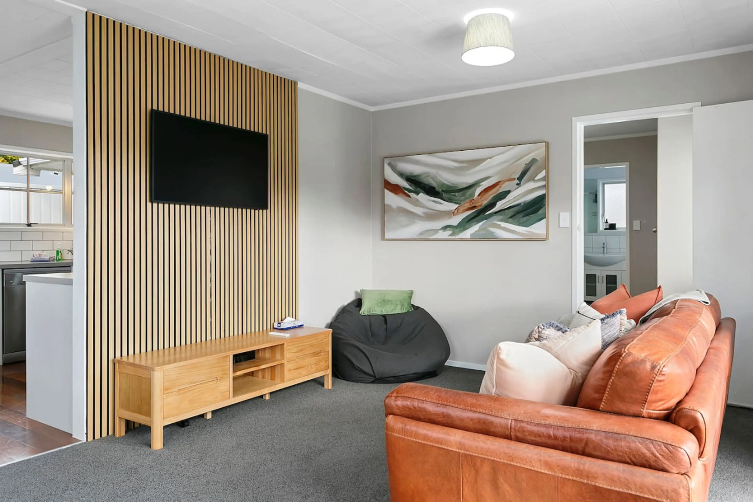 Living room with gray walls, wooden slat accent wall, wall-mounted TV, abstract painting, brown leather sofa with pillows, small black bean bag chair, and visible doorway to bathroom.