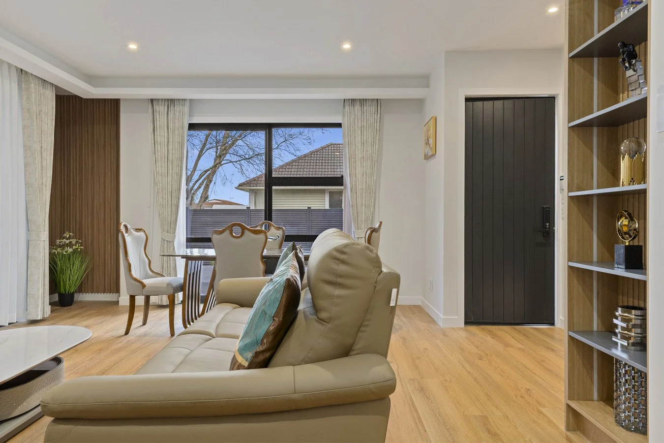 Living room with beige leather sofa, dining table with chairs, large window showing outdoor trees and houses, wooden flooring, and a front door.