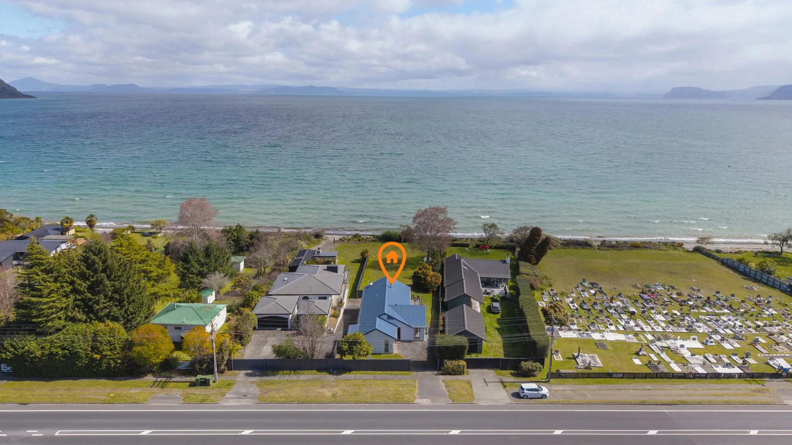 Aerial view of residential houses along a beachfront with a cemetery to the right, ocean, and mountains in the distance.