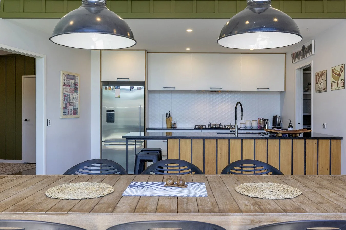 A modern kitchen with white cabinets, a white textured backsplash, and a black countertop on an island with a wooden base. There are three black barstools tucked under the island. The kitchen features a stainless steel refrigerator, a gas stove, and 