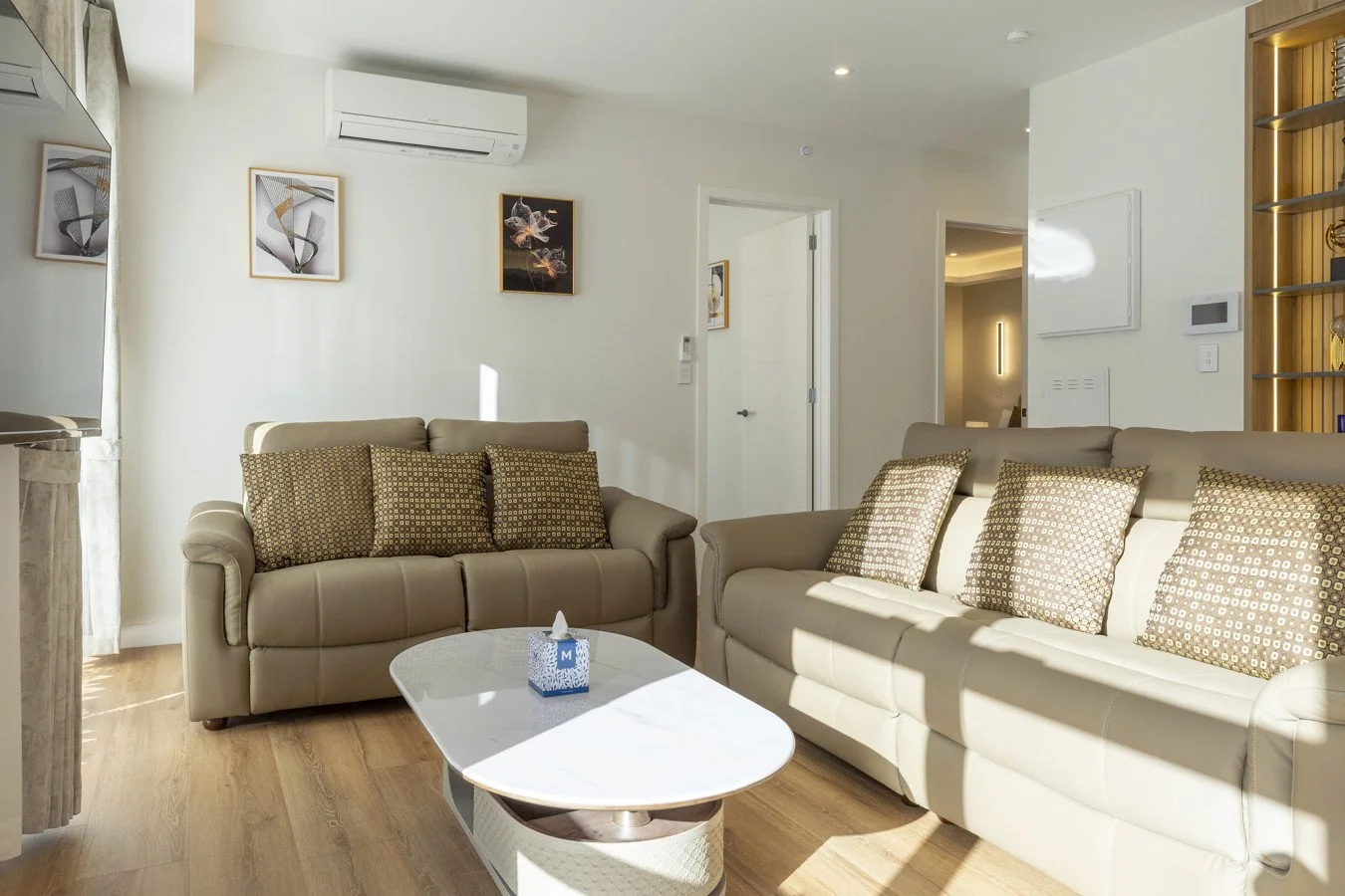 Living room with two beige sofas, patterned cushions, a white coffee table with a tissue box, artwork on the walls, a window with curtains, and a split air conditioning unit.