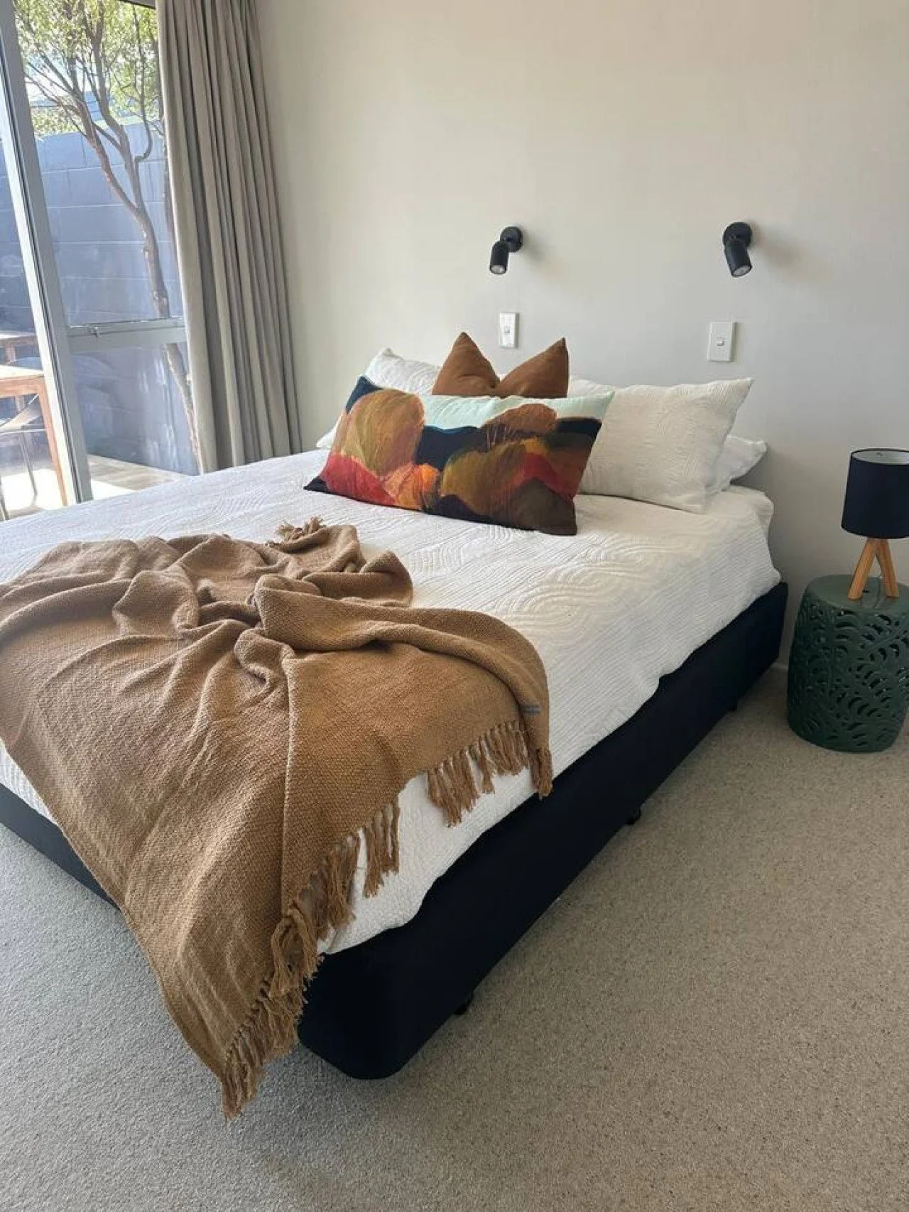 A neatly made bed with a beige throw blanket, white bedsheets, and several pillows including one with a colorful abstract pattern in a modern bedroom. There's a nightstand with a black lamp, a sliding glass door with curtains, and a view of trees out