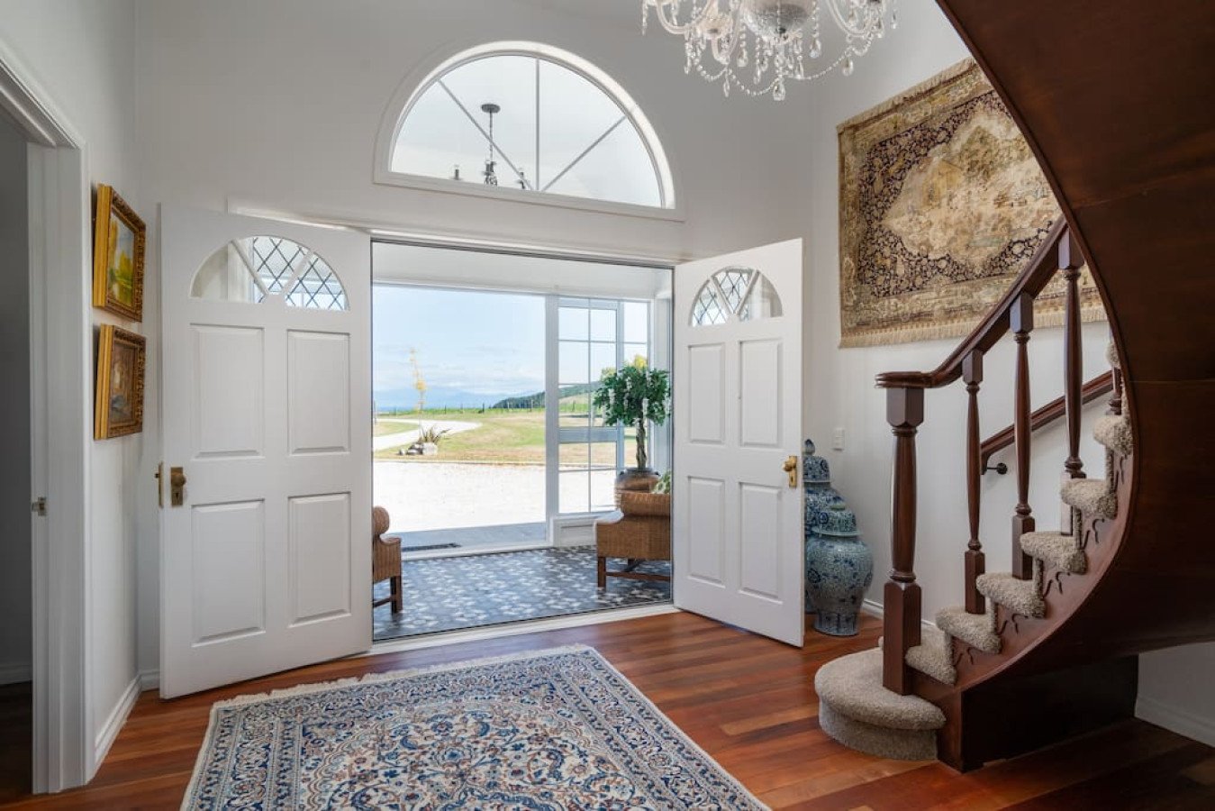 Entrada de casa con puertas dobles blancas, alfombra persa, escaleras de madera y escultura de cerámica en la esquina. Ventanas con vista al campo y un árbol en la entrada.