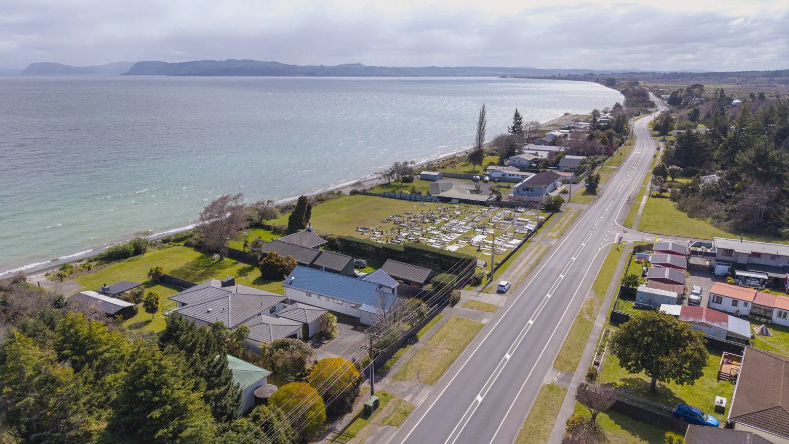 Aerial view of a coastal town with houses, a cemetery, and a road adjacent to the beach, with water and distant landforms in the background.