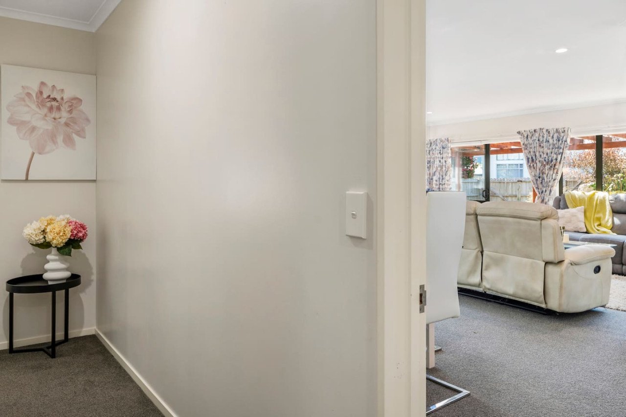 View of a living room with beige sofas, floral curtains, windows with outdoor scenery, and a yellow blanket, seen through an open door from a plain white hallway that has a small table with a flower arrangement and a floral picture on the wall.
