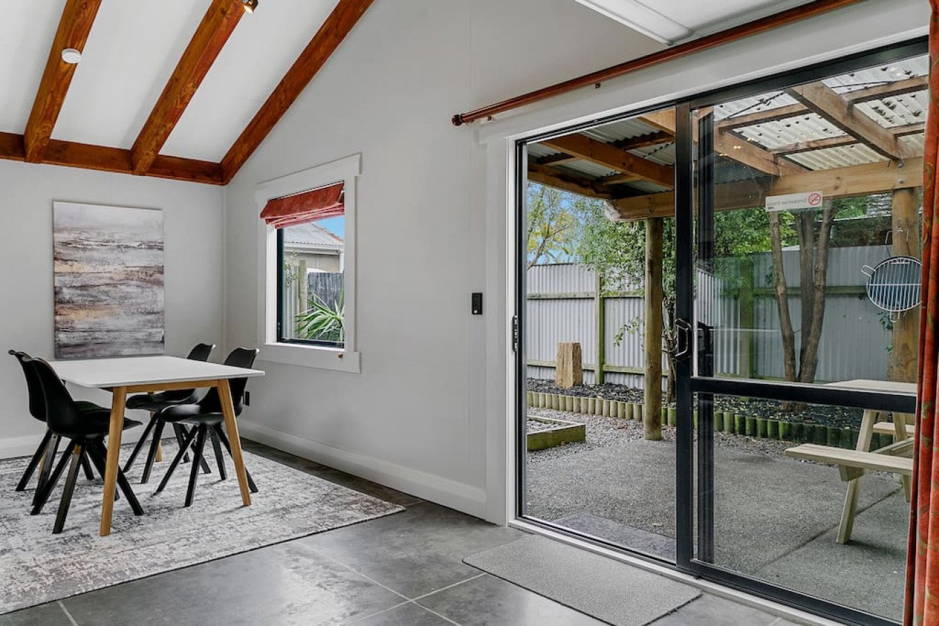 Interior view of a dining room with a white table, black chairs, window with red valance, and sliding glass door leading to a backyard patio with a picnic table, trees, and a fence.