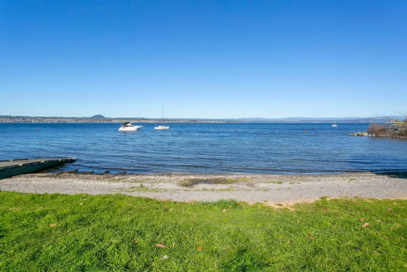 A lakeshore scene with a grassy foreground, a sandy and gravel shoreline, and calm blue water with boats floating. The background includes a clear sky and distant landforms.