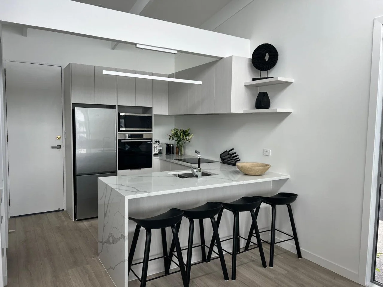 Modern kitchen with white marble island, black bar stools, stainless steel appliances, and minimalist decor.